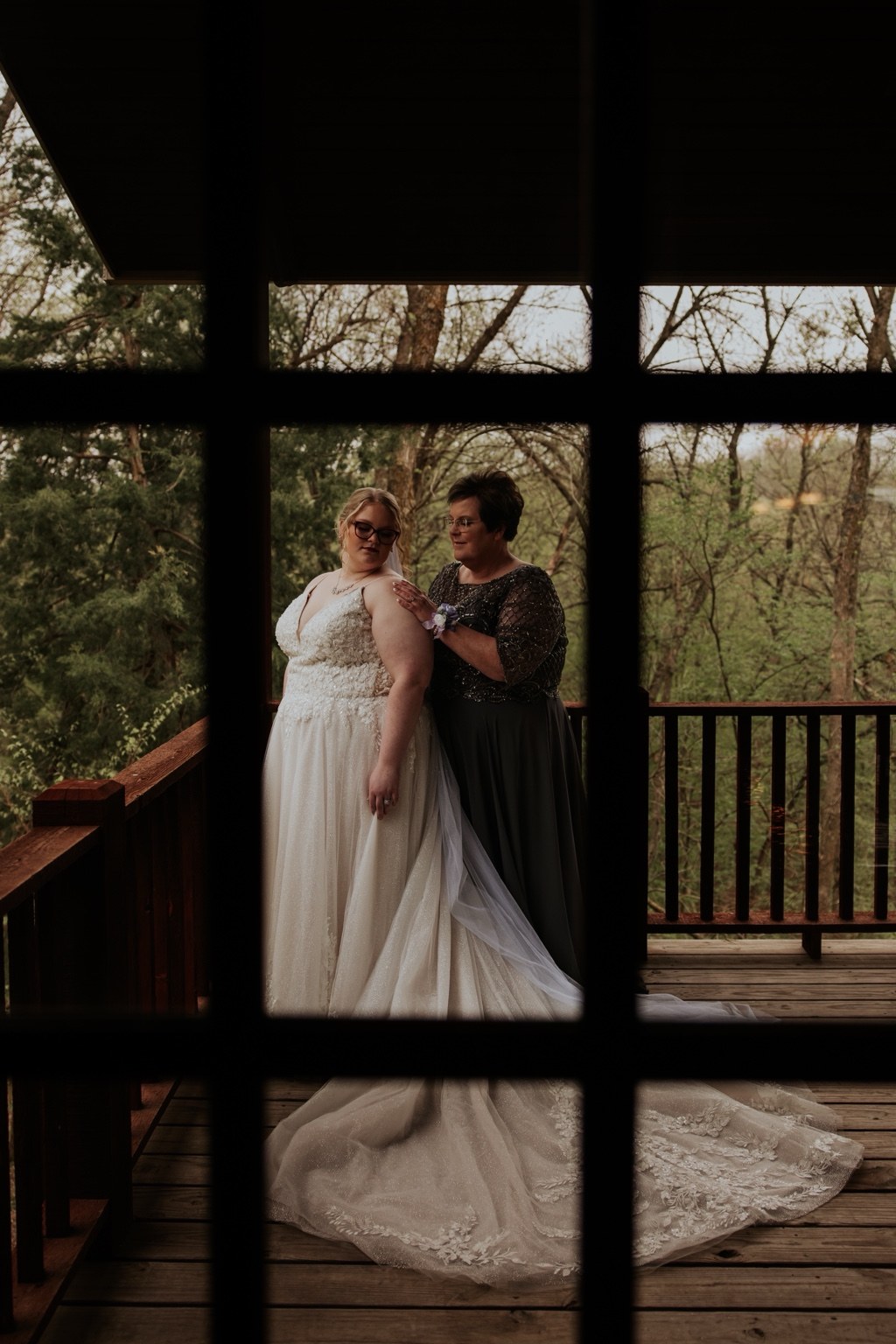 A bride and loved one on a wooden porch framed through a window