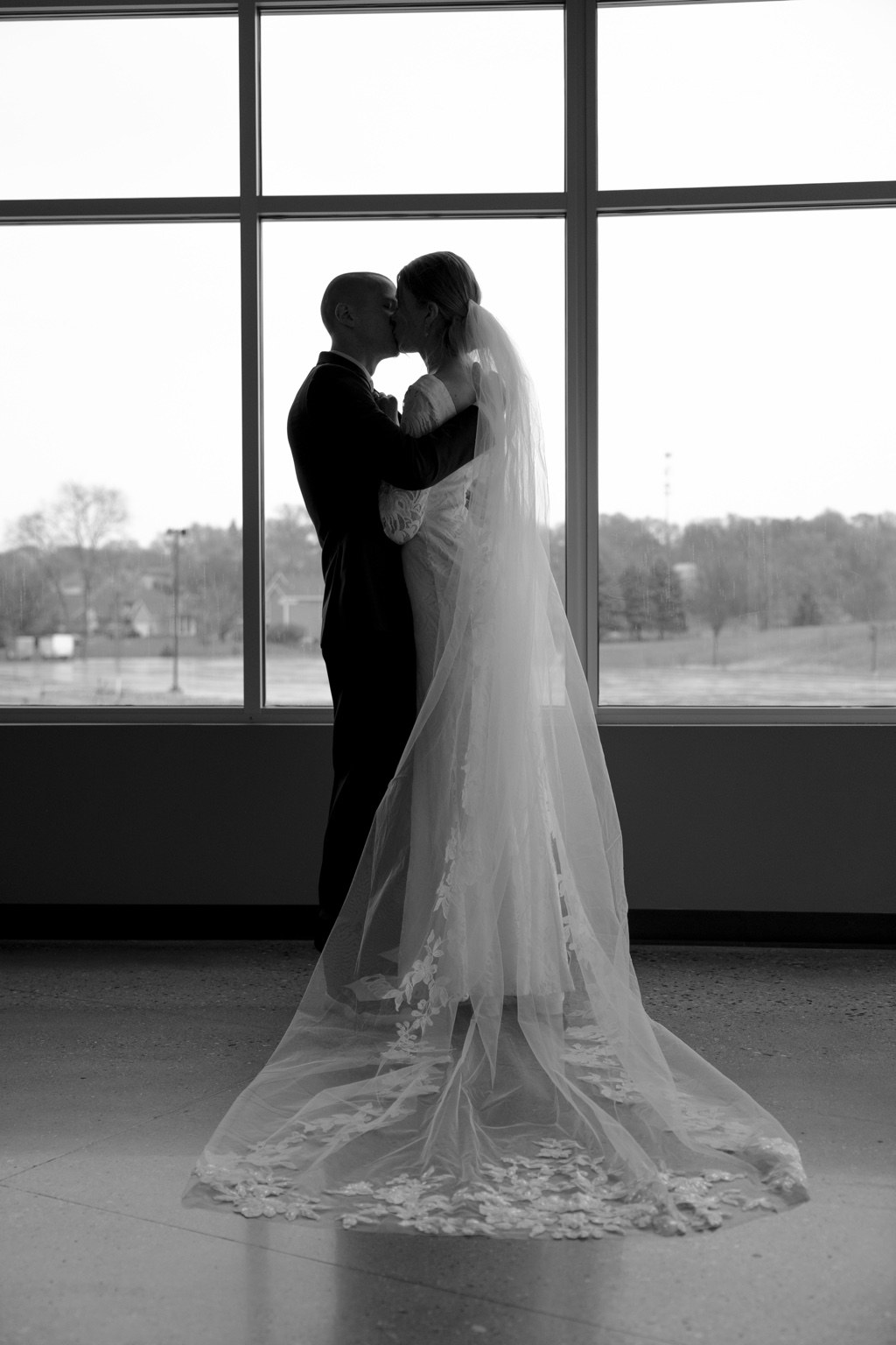 A black and white silhouette of newlyweds kissing by a window